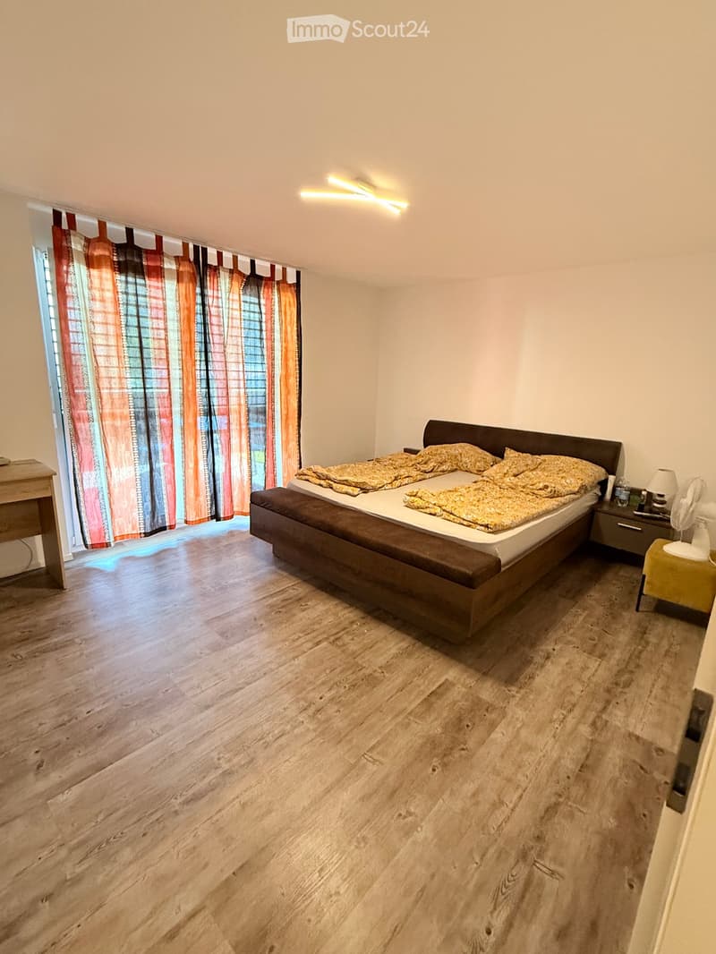 A well-lit bedroom with wooden flooring, a large bed with patterned bedding, a nightstand with a lamp, a small fan, and a colorful curtain covering a window.