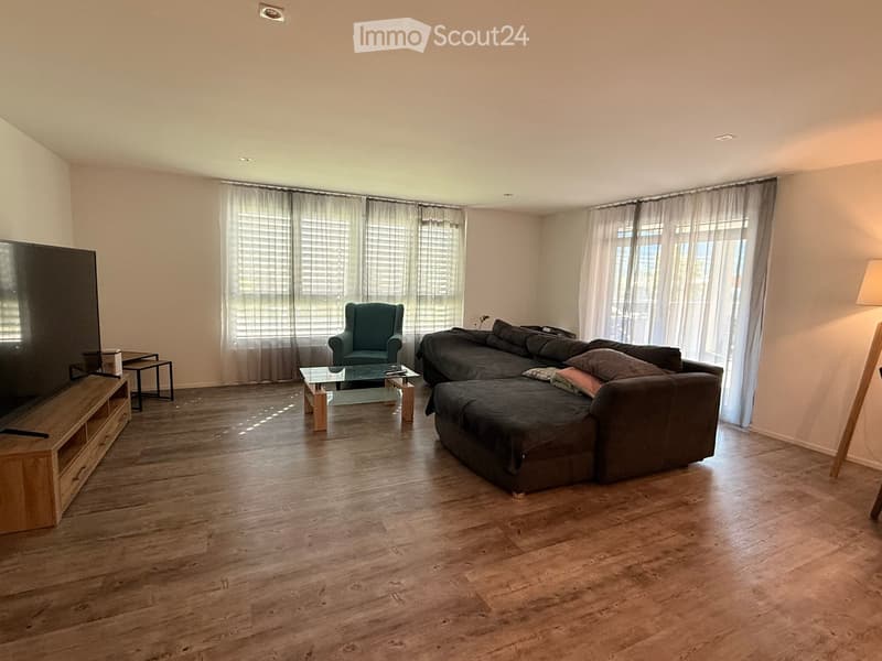 Modern living room with hardwood flooring, large windows with curtains, a sectional sofa, armchair, coffee table, TV stand, and small side table.