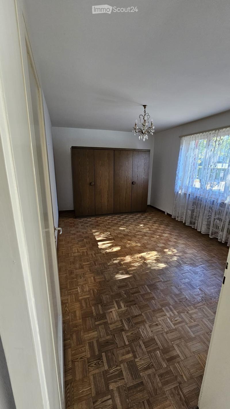 Empty room with wooden herringbone flooring, white walls, a large wooden wardrobe, and a chandelier hanging from the ceiling. Natural light filters through sheer curtains on the window.