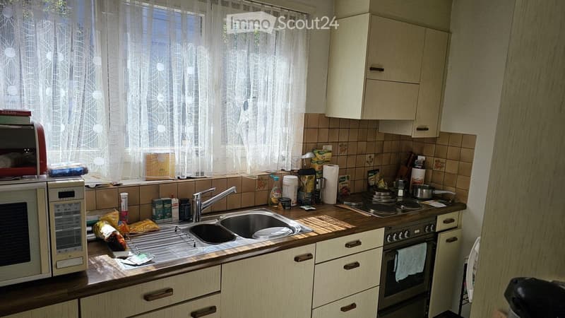 A small kitchen with light wooden cabinets, a double sink, a microwave, and an oven. The counter is cluttered with various items including a toaster, a bottle of dish soap, and some food packages.