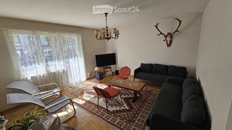 Living room with a dark grey sofa, coffee table, red chairs, a chandelier, and a television on a stand. The room features a patterned rug, wooden flooring, and large windows with sheer curtains.