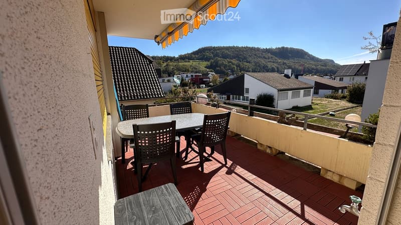 Balcony with a round table and four chairs, red-tiled floor, overlooking a residential area with houses and hills.