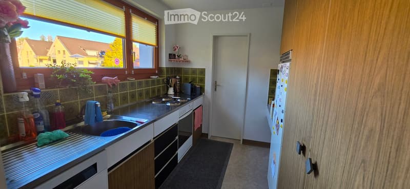 The kitchen features a stainless steel sink, a four-burner stove, a refrigerator, and wooden cabinets. Cleaning supplies and a dish soap bottle are on the counter, and a small shelf holds decorative items. The room has large windows with blinds and a view