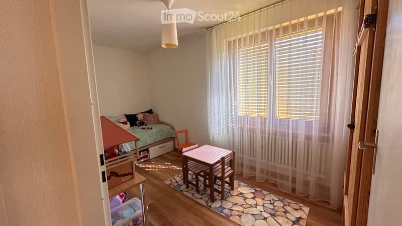 A cozy children's bedroom with a bed, small table and chairs, toy house, and storage unit. The room features wooden flooring, a window with white curtains, and a decorative stone rug.