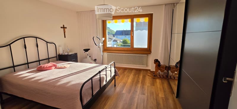 Cozy bedroom with wooden flooring, a metal-framed bed with pink bedding, a nightstand, an exercise bike, a toy horse, and a cross on the wall. Large window with white curtains offers a scenic view.