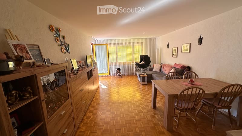A well-lit living room with wooden flooring, a sofa, a dining table with chairs, a cabinet with glass doors, and various decorative items including picture frames and a mirror. There is also a baby stroller and a bicycle near the window.
