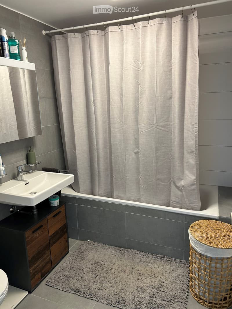 Modern bathroom with a white sink, chrome faucet, bathtub with a grey shower curtain, dark grey tiles, a woven stool, and a grey rug.