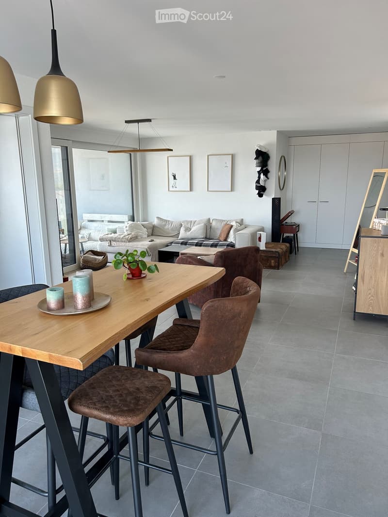 Modern living room with a sofa, coffee table, and dining area featuring a wooden table and brown chairs. The space includes a mirror, a plant, and decorative candles.
