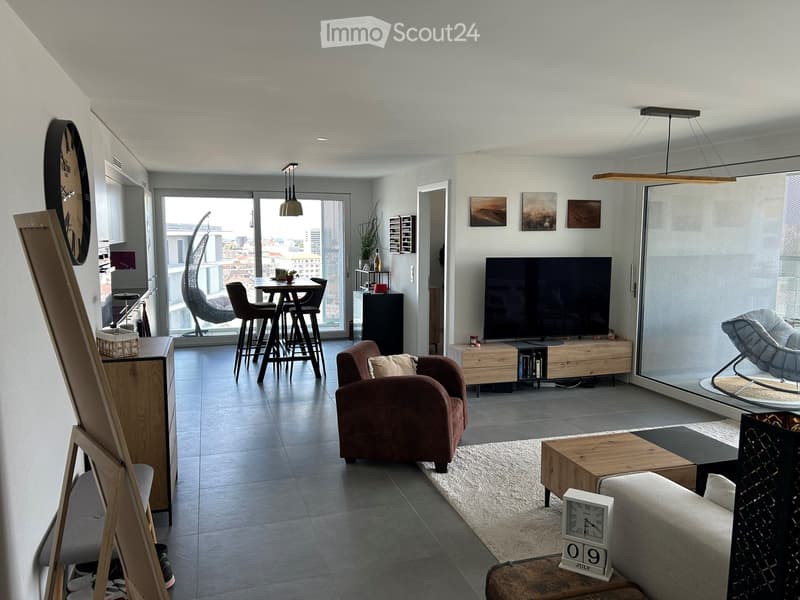 Modern living room with large windows, grey sofa, coffee table, armchair, TV, dining area with chairs and table, and decorative items.