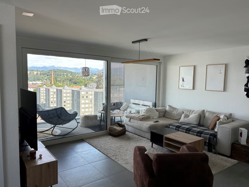 Modern living room with large windows offering a scenic view, featuring a grey sectional sofa, coffee table, wooden side table, hanging pendant lights, and a TV on a stand.