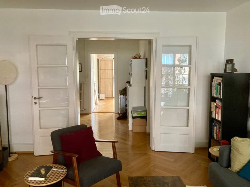 Living room with wooden flooring, a grey sofa, a wooden chair with a red cushion, a small round table, a bookshelf, and a floor lamp.