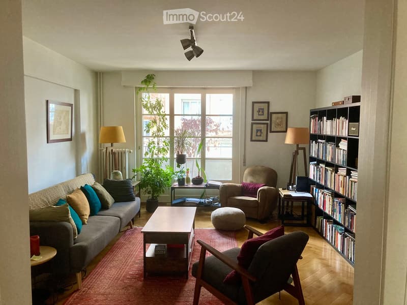 A cozy living room with a grey sofa, wooden coffee table, armchair, bookshelf, and decorative plants. The room features large windows with a view, warm lighting, and a mix of modern and classic furniture.