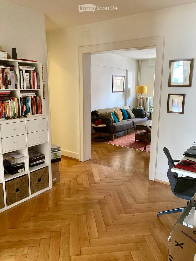 A modern, well-lit living space with a wooden floor, a grey sofa with colorful cushions, a coffee table, a small side table, a bookshelf, and a desk with a chair and laptop.