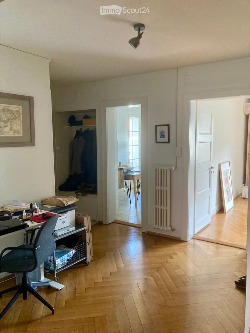 A furnished room with wooden flooring, a desk with a chair, a printer, and a radiator. There is an open doorway leading to another room with a table and stool.