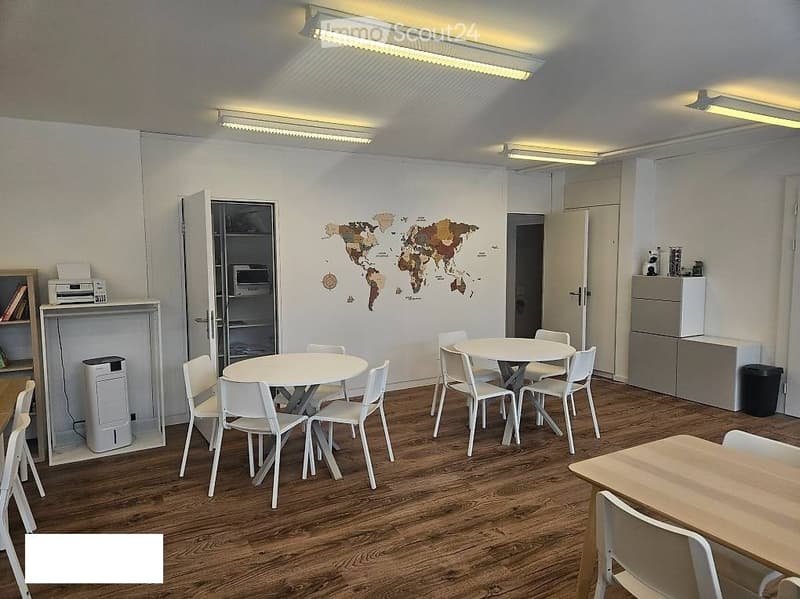 Bright, modern room with wooden flooring, white furniture including tables and chairs, a world map on the wall, a refrigerator, and a shelf with various items.