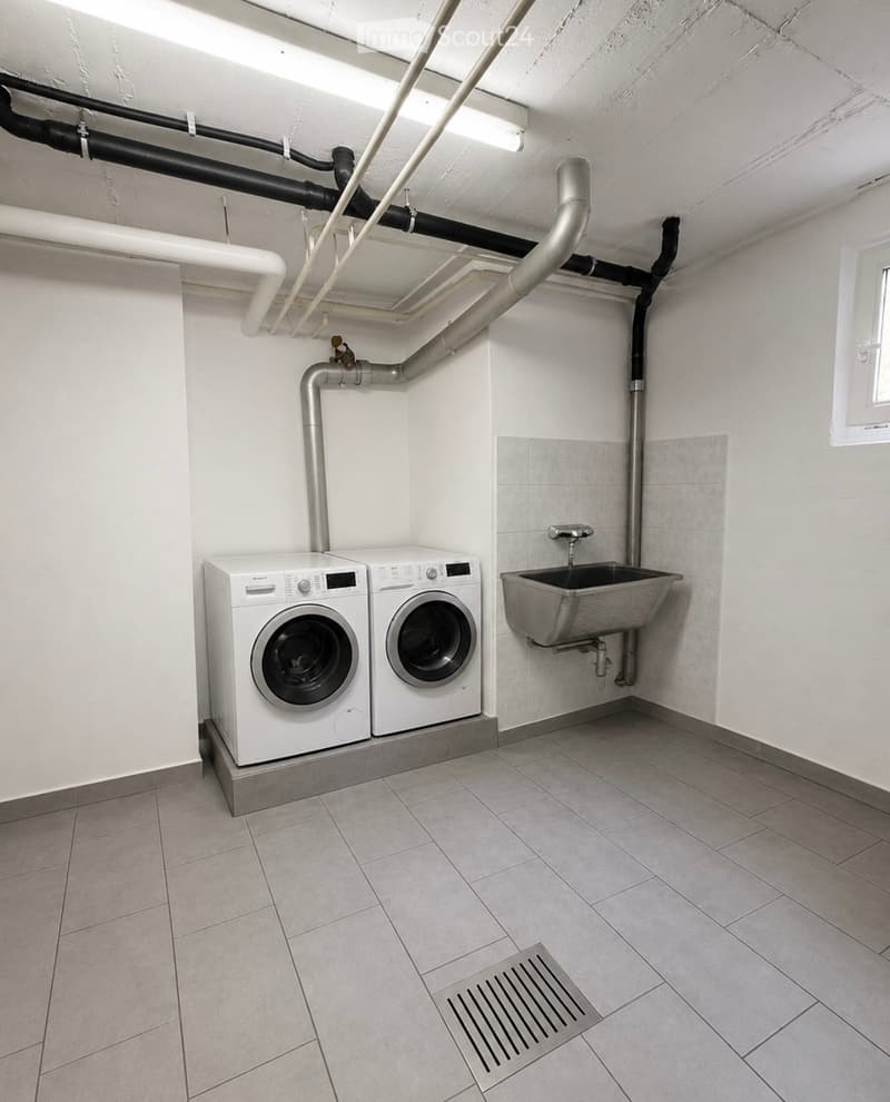 Utility room with tiled floor, white walls, two washing machines, and a sink.