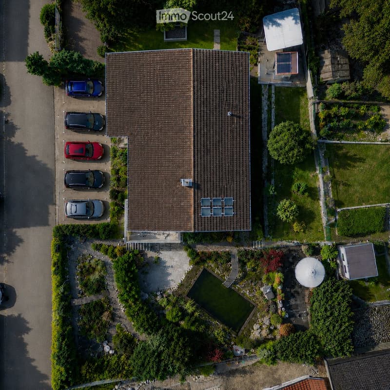 Aerial view of a two-story house with a brown tiled roof, surrounded by a well-maintained garden, a swimming pool, and a parking area with several cars.