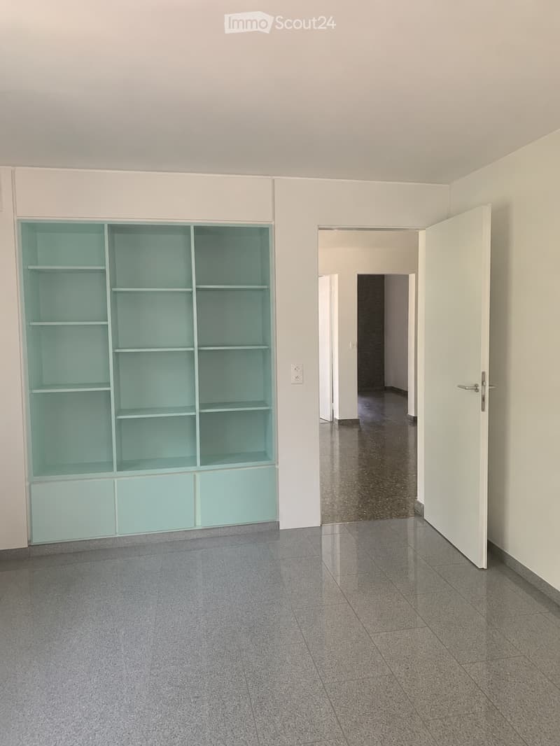 Empty room with grey tiled floor, white walls, built-in turquoise shelving unit, and an open doorway leading to another room.