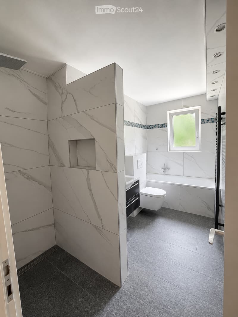 Modern bathroom with marble walls, a bathtub, toilet, sink, and a large mirror. Features a window with frosted glass and a shower area with a glass enclosure.