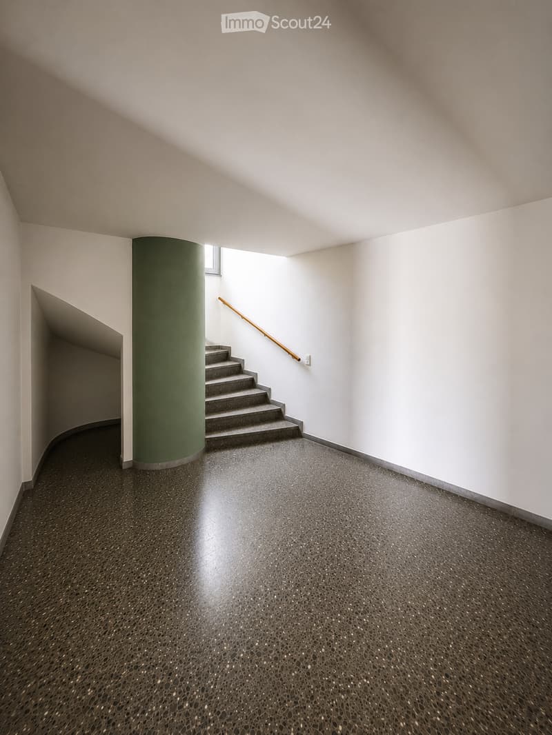Modern hallway with a staircase, green cylindrical column, dark speckled floor, and white walls.