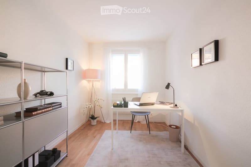 A modern, well-lit room with a wooden floor, white walls, and large windows. It features a desk with a laptop, a chair, a floor lamp, a table lamp, a shelf with books and decorative items, and a small plant in a pot.