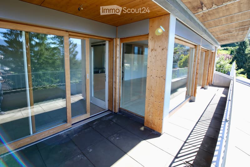 Modern balcony with large glass sliding doors, wooden beams, tiled floor, and a view of greenery.