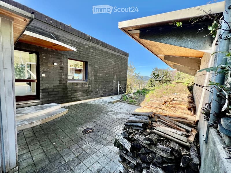 Modern outdoor patio area with stone-tiled flooring, stacked firewood, and a view of a contemporary house with dark brick walls and large windows.