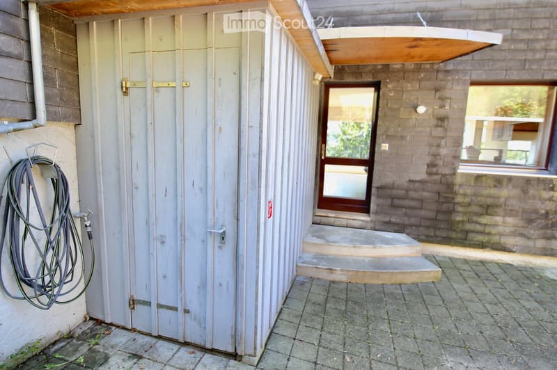 Outdoor area with a small shed, hose reel, steps leading to a door, and a view of a house with brick walls and windows.