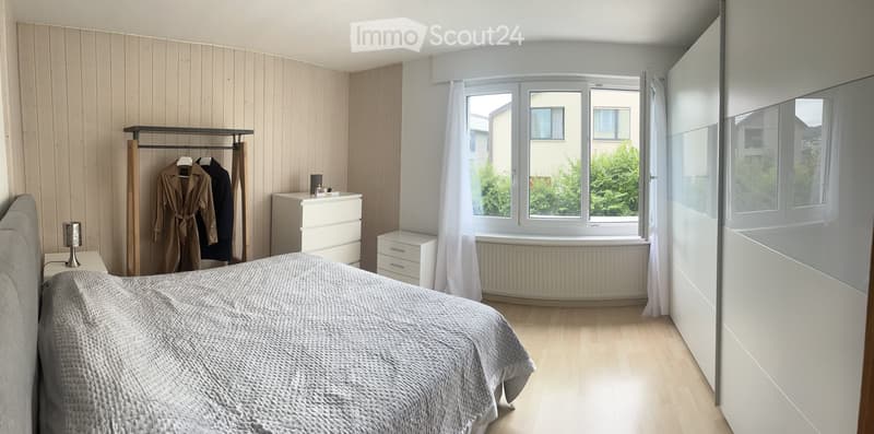 Modern bedroom with light wooden flooring, a large bed with a grey duvet, a nightstand with a lamp, a wardrobe, and a coat rack with coats.