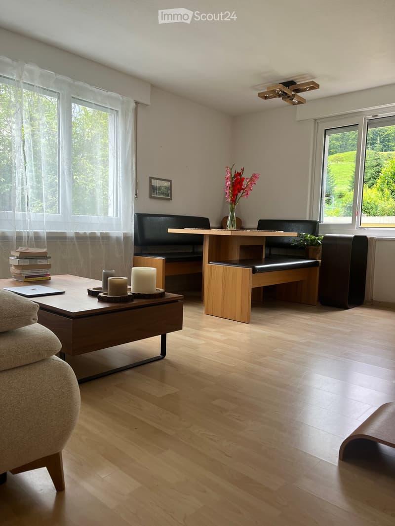 Modern living room with light wooden flooring, large windows, a sofa, coffee table, bench seating, and decorative items including candles and a vase with flowers.