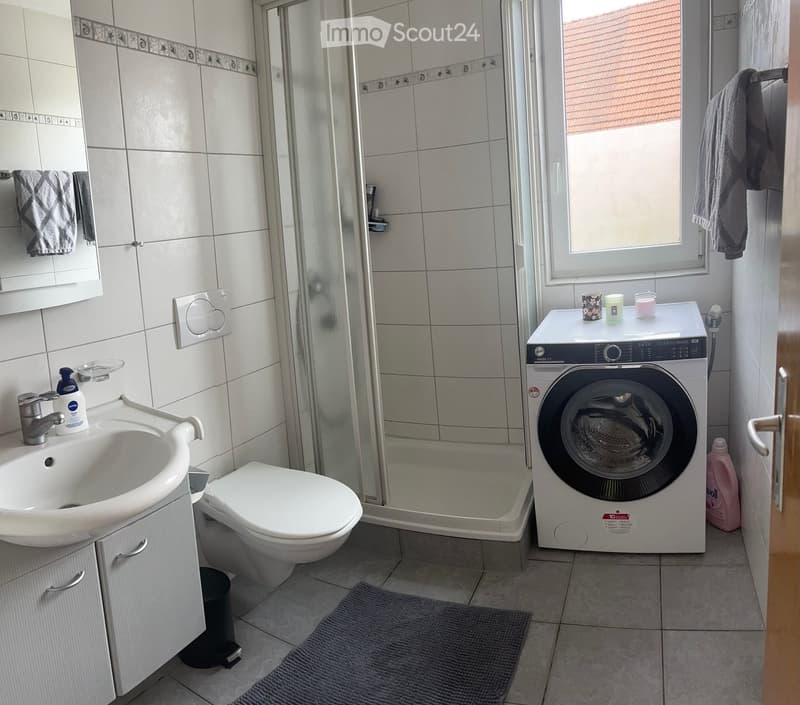 Modern bathroom with white tiles, a sink, toilet, shower, and a washing machine. Includes a mirror, towels, and small decorative items on the washing machine.