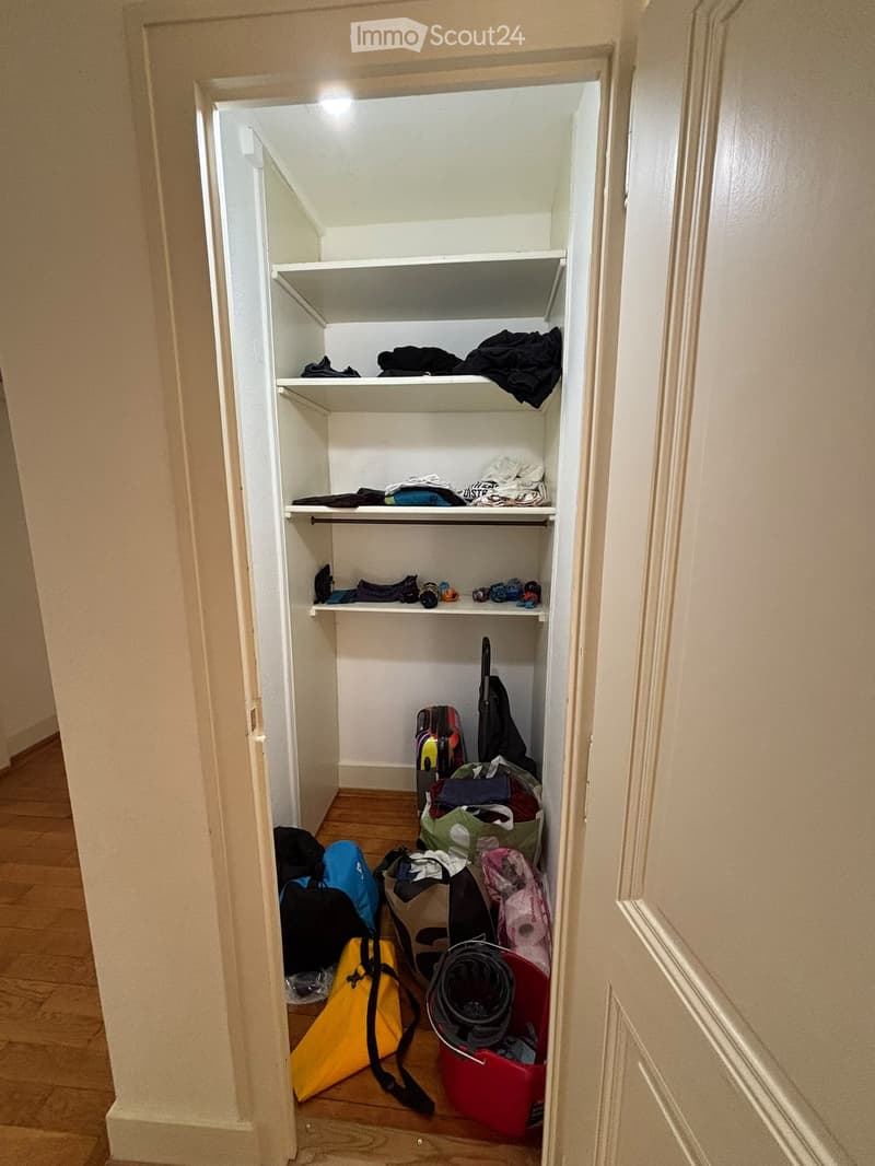A narrow hallway leading to an open closet with wooden shelves and various stored items including bags, clothing, and miscellaneous objects.