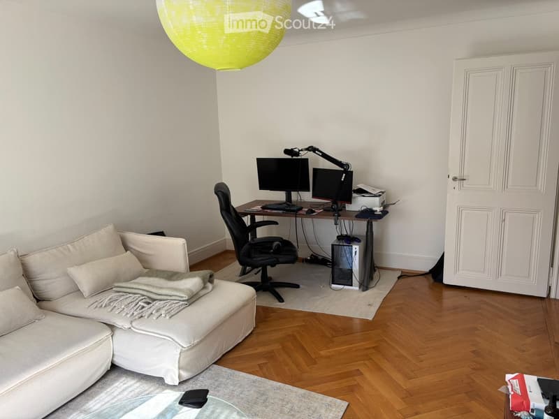A modern living room with a white sofa, a desk with a computer and two monitors, a chair, and a round glass coffee table.