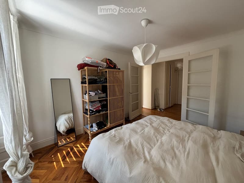Bedroom with wooden floor, large mirror, bed with white bedding, and a wooden shelf with folded clothes and bags.
