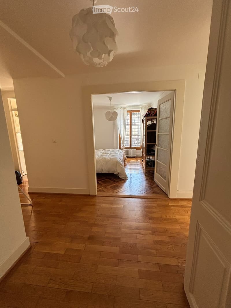 A well-lit hallway with wooden flooring and white walls, leading to a bedroom with a bed, radiator, and shelving unit.
