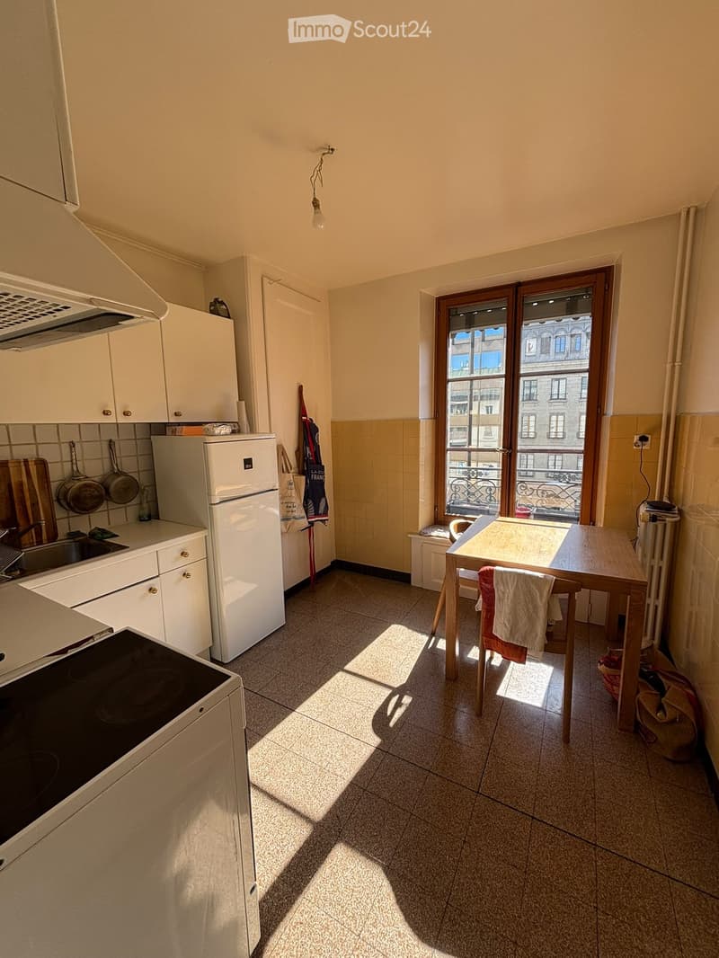 A compact kitchen with white cabinets, a refrigerator, stove, sink, and a small wooden dining table with chairs. The room features tiled floors and a large window with a view of the city.
