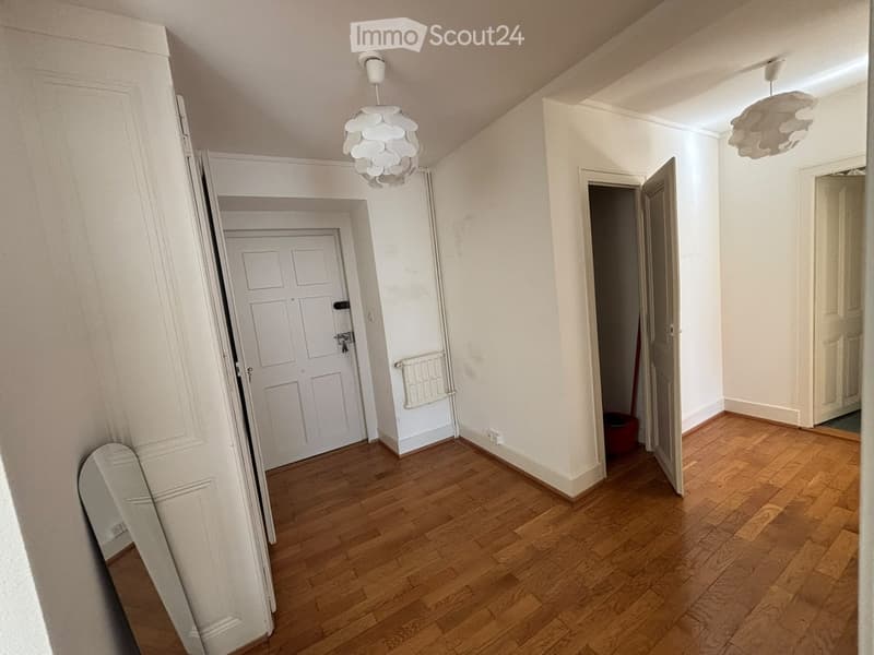 Empty hallway with wooden flooring, white walls, and several doors leading to other rooms. Two modern pendant lights are suspended from the ceiling.