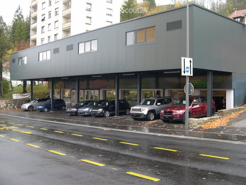 Modern multi-story building with a flat roof and large windows, featuring a covered parking area with several parked cars.