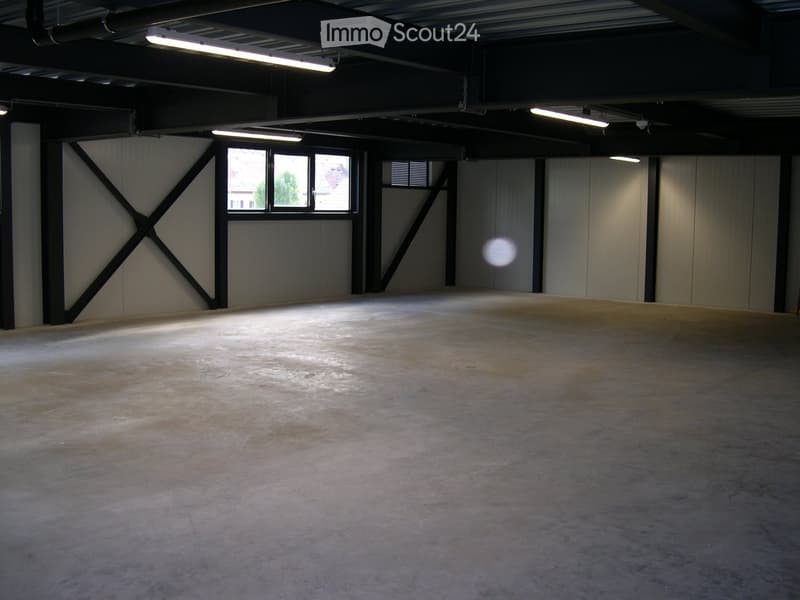 Empty warehouse space with concrete floor, exposed metal beams, and large windows allowing natural light.