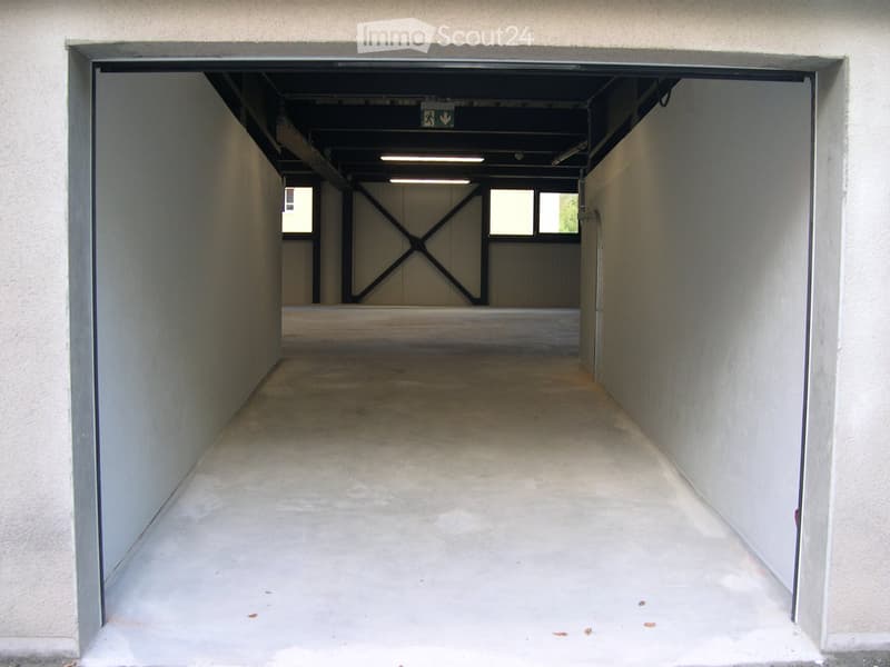 A narrow, concrete-floored corridor with white walls, leading to a parking area with visible metal structural beams and windows at the far end.