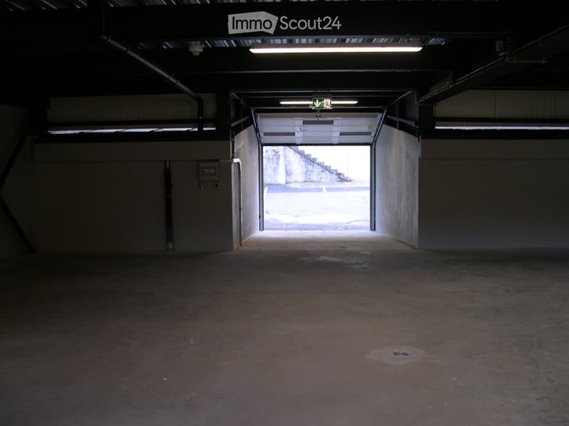 Empty underground parking space with concrete floors and walls, fluorescent lighting, and an open doorway leading outside.
