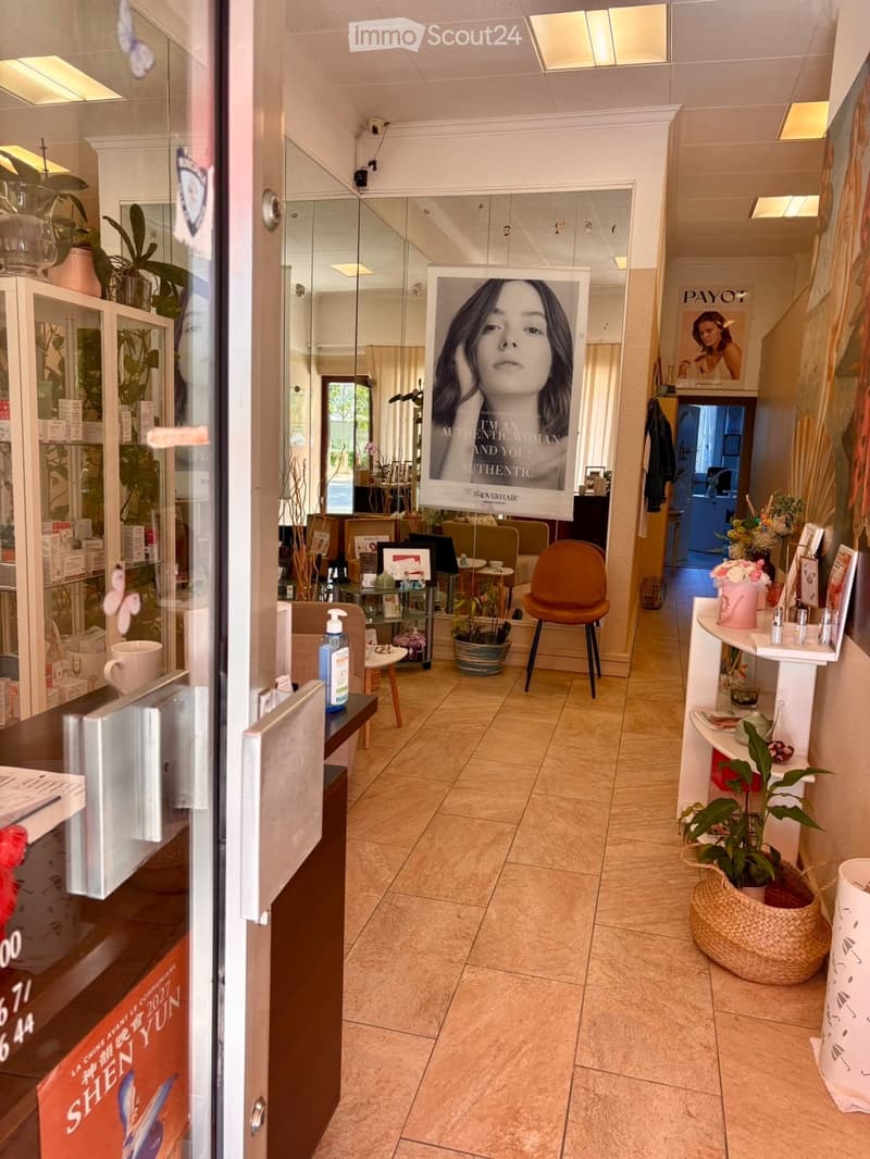 A well-lit hallway inside a beauty salon with a tiled floor, a glass display cabinet, a poster, a potted plant, and a small seating area with a chair and table.
