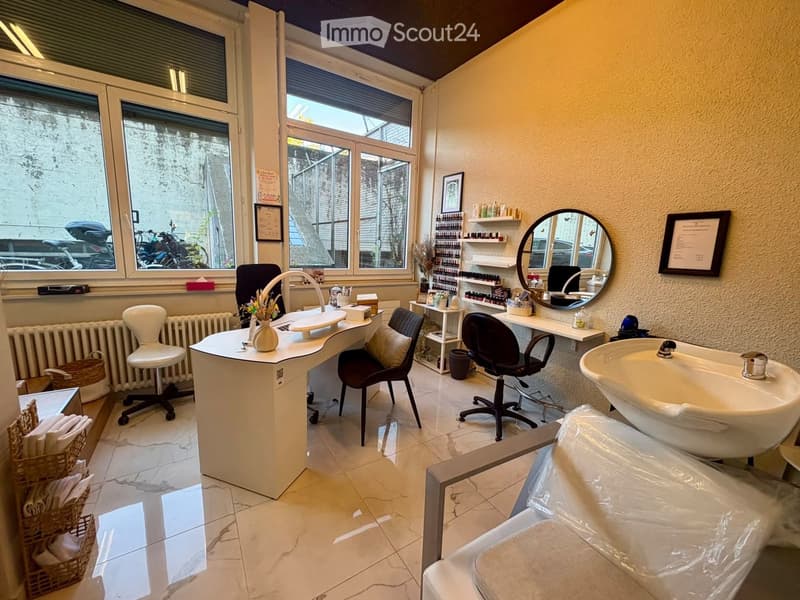 Well-lit salon with marble floors, large windows, a sink, a mirror, a chair, a table, and shelves with various items.