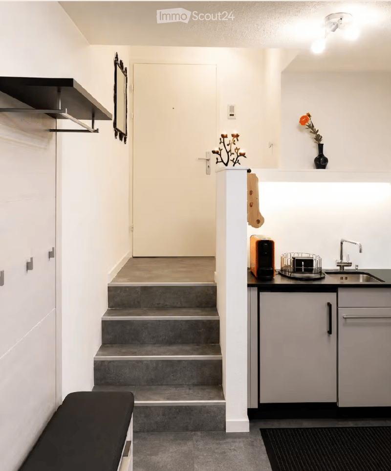 Modern hallway with white walls, grey carpet, a small bench, and a staircase leading to an upper level. A small kitchen area is visible with a sink, faucet, and countertop.