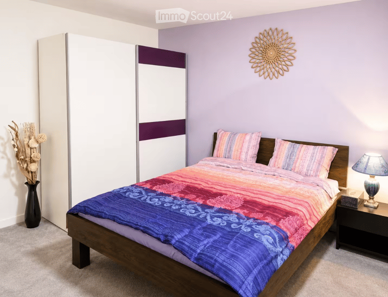 A modern bedroom with a wooden bed, colorful bedding, a nightstand with a lamp, a vase with dried flowers, and a large wardrobe with a purple accent.