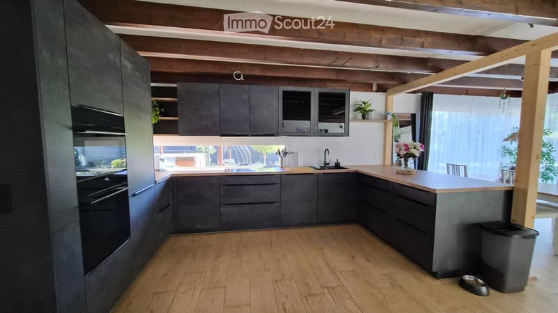 Modern kitchen with dark grey cabinets, wooden countertop, sink, and built-in oven. Features a central island with a vase of flowers and a stool, and a trash bin on the floor.