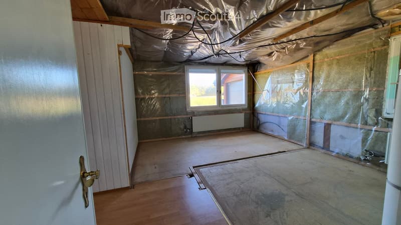 Empty room with exposed wooden beams, insulation, and a window. Bare walls and unfinished flooring.