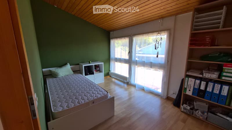 A small, furnished bedroom with a single bed, a nightstand, a wooden shelf, and a dreamcatcher hanging from the ceiling. The room has wooden flooring and large windows with sheer curtains.