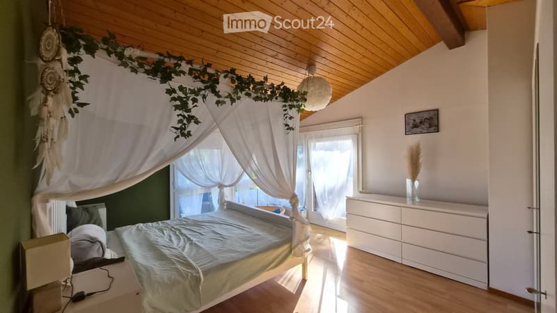 Cozy bedroom with a wooden bed, nightstand, dresser, and a dreamcatcher above the bed. The room features a slanted wooden ceiling and large windows with white curtains.