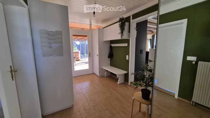 Bright hallway with wooden flooring, white bench, small plant on a stool, and a view to an outdoor area through a glass door.